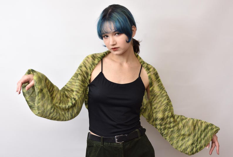 May include: A black spaghetti strap tank top paired with a lime green and dark olive green variegated knit shrug featuring long, wide sleeves. The shrug has a loose, open-front design that drapes across the shoulders and arms, showcasing a textured knit pattern.