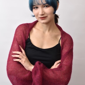 May include: A deep burgundy, sheer, knitted shrug featuring long, voluminous sleeves with a loose, airy texture. The garment is worn over a simple black camisole, showcasing a lightweight, open-knit construction that drapes softly around the shoulders.