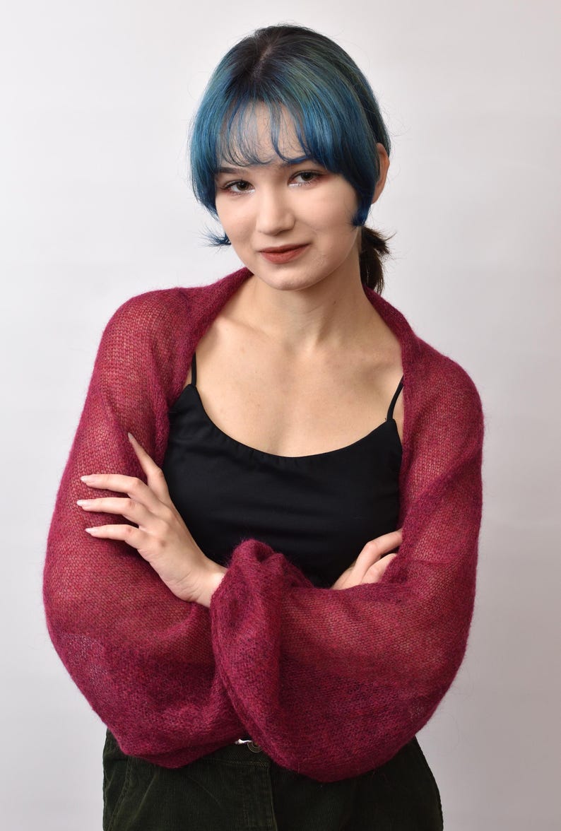 May include: A deep burgundy, sheer, knitted shrug featuring long, voluminous sleeves with a loose, airy texture. The garment is worn over a simple black camisole, showcasing a lightweight, open-knit construction that drapes softly around the shoulders.