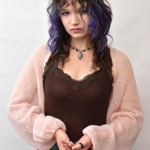 May include: A light pink, sheer, long-sleeved knit bolero cardigan worn over a dark brown camisole with lace trim. The bolero features a loose, airy texture with cuffed sleeves, paired with a multi-colored beaded necklace and a teal and brown skirt.