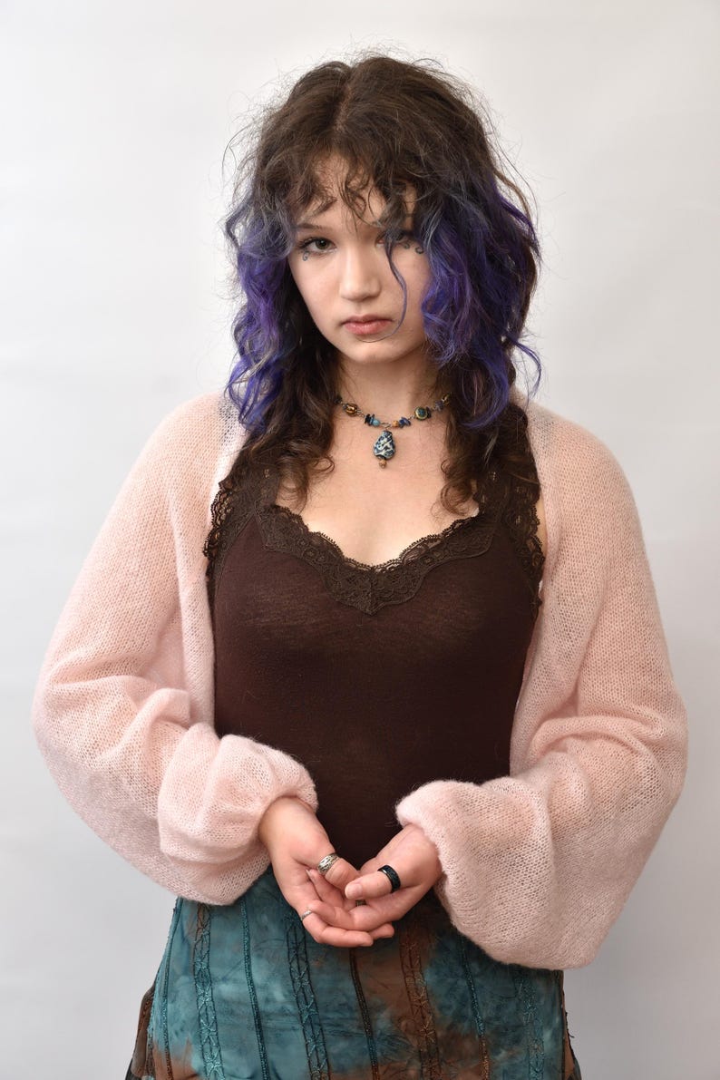 May include: A light pink, sheer, long-sleeved knit bolero cardigan worn over a dark brown camisole with lace trim. The bolero features a loose, airy texture with cuffed sleeves, paired with a multi-colored beaded necklace and a teal and brown skirt.