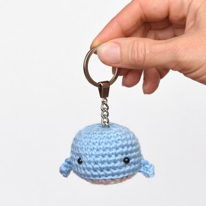 May include: A light blue crocheted whale keychain with black eyes. The whale is attached to a silver key ring and chain.