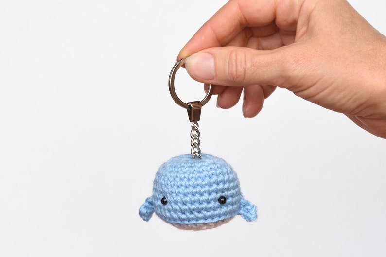 May include: A light blue crocheted whale keychain with black eyes. The whale is attached to a silver key ring and chain.