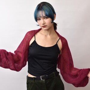 May include: A deep burgundy, sheer, knitted shrug featuring long, voluminous balloon sleeves. The garment is worn over a black camisole top and dark trousers with a black belt. The knit texture is lightweight and semi-transparent.