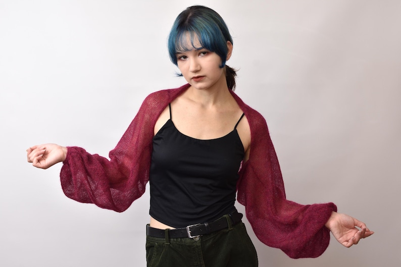 May include: A deep burgundy, sheer, knitted shrug featuring long, voluminous balloon sleeves. The garment is worn over a black camisole top and dark trousers with a black belt. The knit texture is lightweight and semi-transparent.