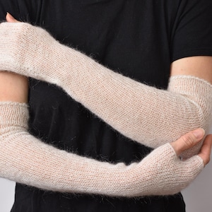 Può includere: Un paio di lunghi scaldabraccia beige senza dita, realizzati in un materiale lavorato a maglia morbido e soffice. Questi scaldabraccia si estendono dalle nocche fino alla parte superiore del braccio, con una trama semplice e leggera.