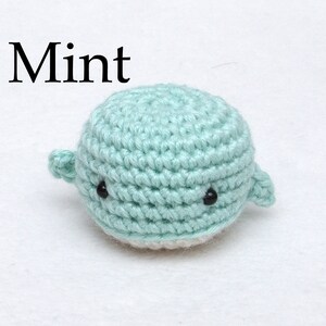 May include: A small crocheted whale in a mint green color with black eyes. The whale is sitting on a white surface. The word "Mint" is written above the whale.