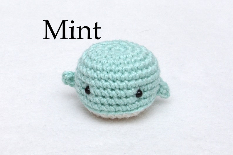 May include: A small crocheted whale in a mint green color with black eyes. The whale is sitting on a white surface. The word "Mint" is written above the whale.