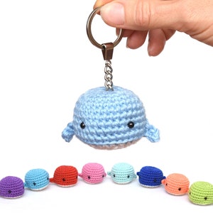 May include: A blue crocheted whale keychain with a silver key ring. The whale is holding a small silver chain. There are nine crocheted whales in a row, each a different color: purple, blue, red, pink, light blue, dark blue, orange, green, and light green.