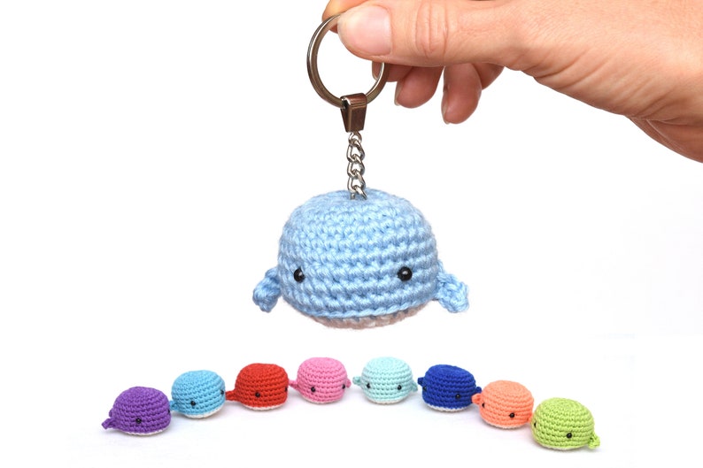 May include: A blue crocheted whale keychain with a silver key ring. The whale is holding a small silver chain. There are nine crocheted whales in a row, each a different color: purple, blue, red, pink, light blue, dark blue, orange, green, and light green.