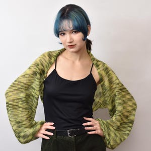 May include: A black camisole top paired with a long-sleeved, open-front bolero shrug featuring a variegated knit pattern in shades of lime green and dark olive. The bolero has loose, flowing sleeves and is worn over a solid black tank top.