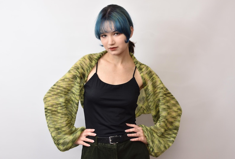 May include: A black camisole top paired with a long-sleeved, open-front bolero shrug featuring a variegated knit pattern in shades of lime green and dark olive. The bolero has loose, flowing sleeves and is worn over a solid black tank top.