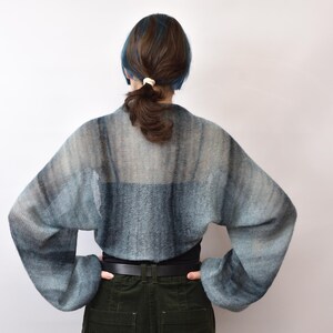May include: A cropped, long-sleeved knit shrug featuring a gradient pattern of muted blue, grey, and charcoal tones. The garment has a loose, airy weave with wide sleeves and a rounded back silhouette, worn over a solid black top and dark trousers.