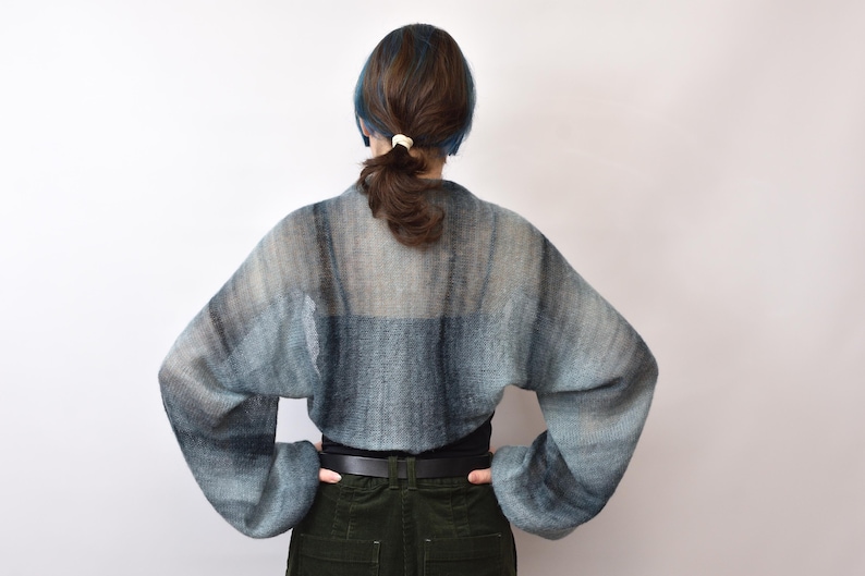 May include: A cropped, long-sleeved knit shrug featuring a gradient pattern of muted blue, grey, and charcoal tones. The garment has a loose, airy weave with wide sleeves and a rounded back silhouette, worn over a solid black top and dark trousers.