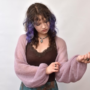 May include: A sheer, light purple bolero shrug with long, voluminous balloon sleeves made of a soft, fuzzy knit fabric. The garment features a wide, open-front design that drapes across the shoulders and arms, showcasing a delicate, semi-transparent texture.