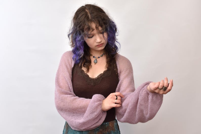 May include: A sheer, light purple bolero shrug with long, voluminous balloon sleeves made of a soft, fuzzy knit fabric. The garment features a wide, open-front design that drapes across the shoulders and arms, showcasing a delicate, semi-transparent texture.