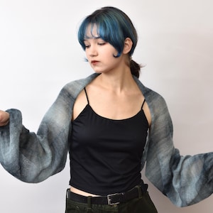 May include: A sheer, blue and gray shrug with long sleeves. The open-front bolero-style top is worn over a black tank top. The sleeves have a gradient effect, transitioning from light to dark blue.