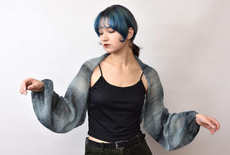 May include: A sheer, long-sleeved shrug featuring a gradient pattern of muted blue, gray, and white tones. The garment has a lightweight, semi-transparent texture with loose, flowing sleeves that extend from the shoulders, worn over a black camisole.