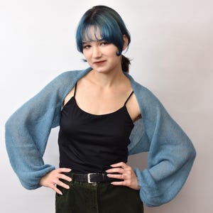 May include: A light blue bolero-style sweater with long sleeves. The sweater is worn over a black tank top and paired with green corduroy pants. The model is standing in front of a white background.