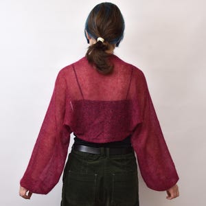 May include: A sheer, deep magenta bolero shrug featuring long, loose-fitting sleeves and a lightweight, open-knit texture. The garment is worn over a dark-colored camisole, dark trousers with a black belt, and has a cropped silhouette.