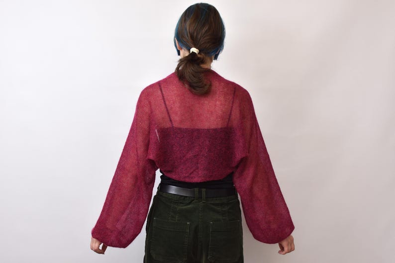 May include: A sheer, deep magenta bolero shrug featuring long, loose-fitting sleeves and a lightweight, open-knit texture. The garment is worn over a dark-colored camisole, dark trousers with a black belt, and has a cropped silhouette.