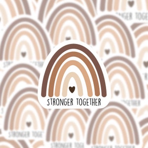May include: A sticker with a brown, beige, and cream rainbow with a small black heart at the center. The text "Stronger Together" is printed below the rainbow.