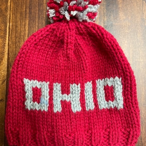 May include: Red knitted beanie with a white and gray pom pom on top. The beanie has the word "OHIO" knitted in gray on the front.