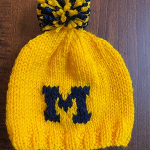 May include: Yellow knitted beanie with a blue and yellow pom pom on top and a blue "M" embroidered on the front.