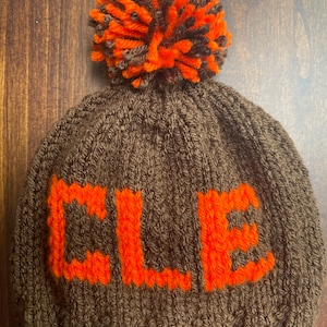 May include: A brown knitted beanie with a large orange and brown pom pom on top. The beanie has the word "CLE" knitted in orange on the front.