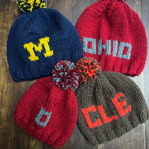 May include: Four knitted beanies with pom-poms. One navy blue with a yellow M, one red with gray OHIO text, one red with a gray D, and one brown with orange CLE text. Each hat features a textured knit pattern and a multi-colored yarn pom-pom on top.