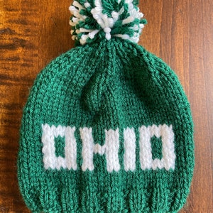 May include: Green knitted beanie with a white pom pom and the word "OHIO" knitted in white on the front.