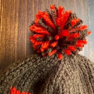 May include: A brown knitted beanie with a large orange and brown pom pom on top. The beanie has a knit pattern and a small orange stripe around the bottom.