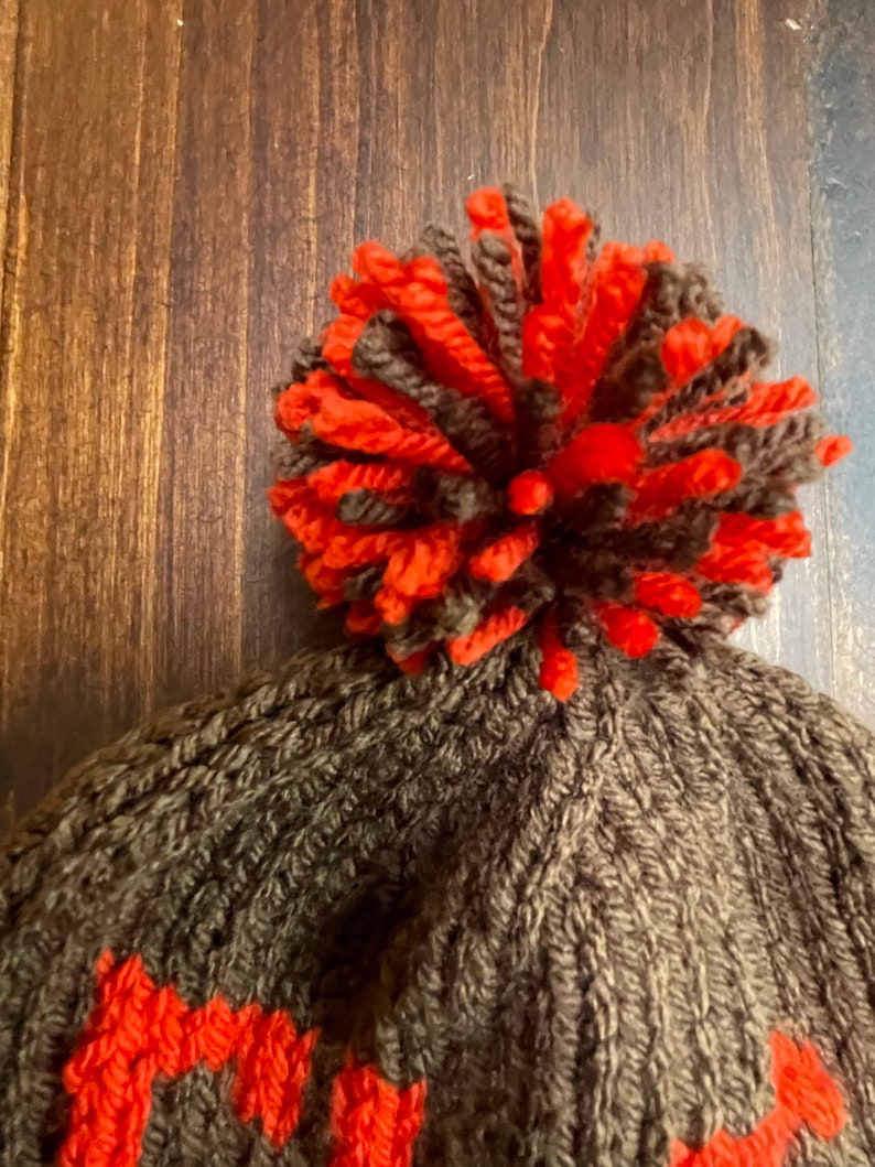 May include: A brown knitted beanie with a large orange and brown pom pom on top. The beanie has a knit pattern and a small orange stripe around the bottom.