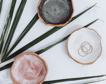 May include: Three small, round, handmade ceramic dishes with uneven, textured edges. One dish is dark grey-green, one is solid white containing three thin gold-toned metal rings, and one is a mottled dusty pink. The dishes have a speckled clay base.
