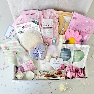 K&ouml;nnte beinhalten: Eine Geschenkbox mit Pflegeprodukten. Enth&auml;lt eine Gesichtsmaske, Seife, einen herzf&ouml;rmigen Keks mit dem Text "You Are Amazing", einen Galaxy-Schokoriegel und Socken mit Blumenmuster. Die Box ist mit rosa und wei&szlig;en Elementen dekoriert.