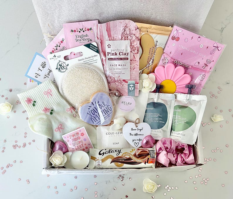 K&ouml;nnte beinhalten: Eine Geschenkbox mit Pflegeprodukten. Enth&auml;lt eine Gesichtsmaske, Seife, einen herzf&ouml;rmigen Keks mit dem Text "You Are Amazing", einen Galaxy-Schokoriegel und Socken mit Blumenmuster. Die Box ist mit rosa und wei&szlig;en Elementen dekoriert.