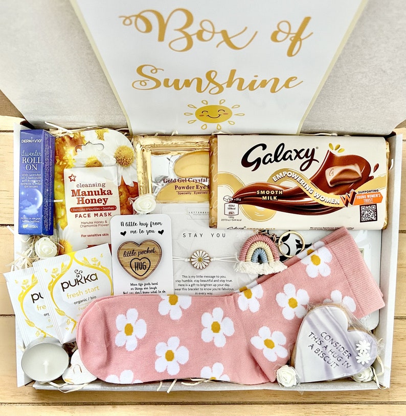 Pu&ograve; includere: Una scatola regalo rosa e bianca piena di vari articoli, tra cui una tavoletta di cioccolato, un piccolo cuore di legno con le parole "little pocket hug", un calzino rosa con margherite bianche, una candela e un piccolo biscotto bianco con le parole "Consider this a hug in a biscuit".