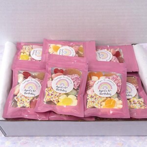 K&ouml;nnte beinhalten: Eine wei&szlig;e Pappschachtel gef&uuml;llt mit mehreren rosa wiederverschlie&szlig;baren Beuteln, die verschiedene Fruchtgummis enthalten. Jeder Beutel hat einen runden Aufkleber mit einem Regenbogenmotiv und dem Text "Thank you for celebrating April's 6th Birthday."