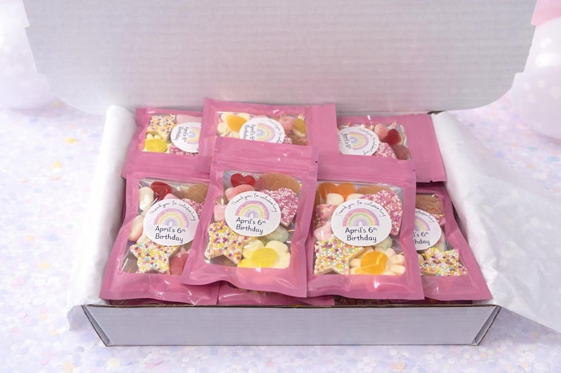 K&ouml;nnte beinhalten: Eine wei&szlig;e Pappschachtel gef&uuml;llt mit mehreren rosa wiederverschlie&szlig;baren Beuteln, die verschiedene Fruchtgummis enthalten. Jeder Beutel hat einen runden Aufkleber mit einem Regenbogenmotiv und dem Text "Thank you for celebrating April's 6th Birthday."