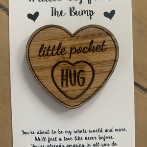 May include: A white card with black text that reads "A little hug from The Bump" with a small black heart on either side of the text. A wooden heart-shaped ornament with the text "little pocket hug" is attached to the card.