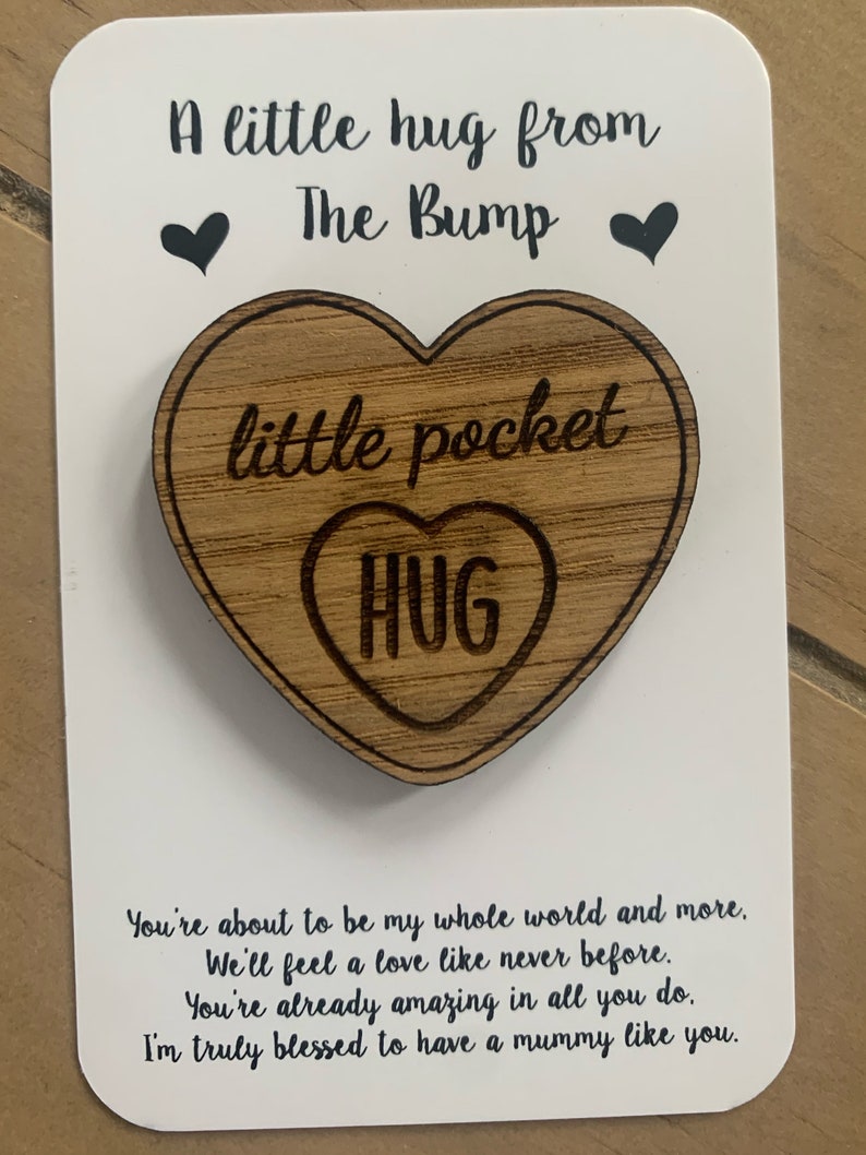 May include: A white card with black text that reads "A little hug from The Bump" with a small black heart on either side of the text. A wooden heart-shaped ornament with the text "little pocket hug" is attached to the card.