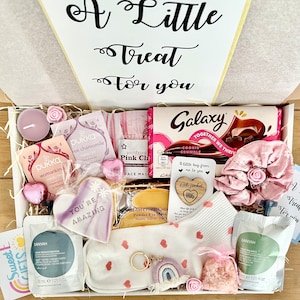 May include: A gift box filled with various items, including a chocolate bar, face masks, tea bags, a sleep mask, and a decorative cookie. The box is decorated with pink and white accents, and the text "A Little Treat for you" is visible.