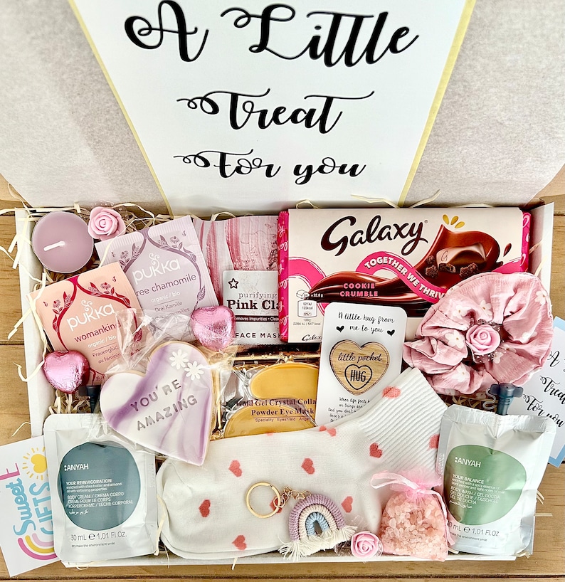 May include: A gift box filled with various items, including a chocolate bar, face masks, tea bags, a sleep mask, and a decorative cookie. The box is decorated with pink and white accents, and the text "A Little Treat for you" is visible.