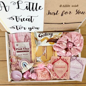 May include: A gift box filled with self-care items. Includes a face mask, Galaxy chocolate bar, eye masks, a scrunchie, tea bags, a candle, a keychain, and a bag of pink crystals. The box has the text "A Little Treat For You" and "A little wish Just for You".