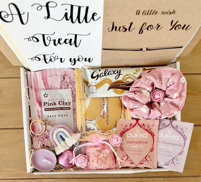 May include: A gift box filled with self-care items. Includes a face mask, Galaxy chocolate bar, eye masks, a scrunchie, tea bags, a candle, a keychain, and a bag of pink crystals. The box has the text "A Little Treat For You" and "A little wish Just for You".