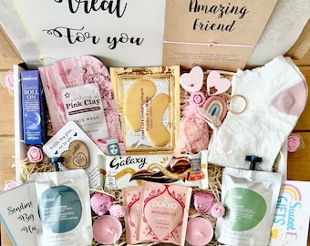 Puede incluir: Una caja de regalo llena de artículos de cuidado personal, que incluyen mascarillas faciales, mascarillas para los ojos, una barra de chocolate, té y un par de calcetines. La caja está decorada con detalles en rosa, incluyendo caramelos en forma de corazón y pequeñas flores. El texto en la caja dice "Treat For You" y "A little wish for an Amazing Friend."