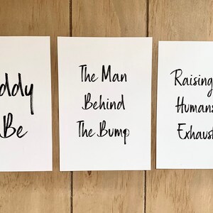 Puede incluir: Tres tarjetas blancas con texto en caligraf&iacute;a negra sobre un fondo de madera. La primera tarjeta dice "Daddy To Be". La segunda tarjeta dice "The Man Behind The Bump". La tercera tarjeta dice "Raising Tiny Humans Is Exhausting".