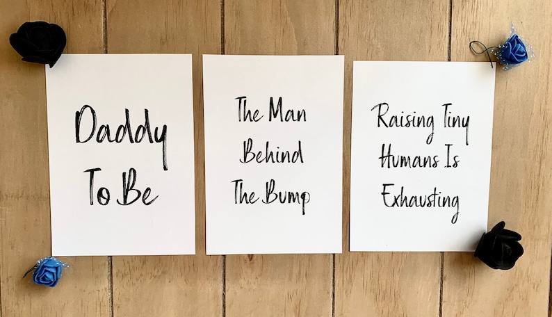 Puede incluir: Tres tarjetas blancas con texto en caligraf&iacute;a negra sobre un fondo de madera. La primera tarjeta dice "Daddy To Be". La segunda tarjeta dice "The Man Behind The Bump". La tercera tarjeta dice "Raising Tiny Humans Is Exhausting".
