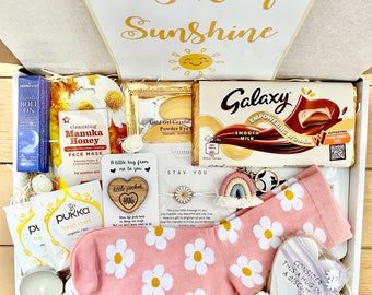 Può includere: Una scatola regalo bianca piena di vari articoli, tra cui una tavoletta di cioccolato, una maschera per il viso, un piccolo cuore di legno con le parole "little pocket hug", un paio di calzini rosa con margherite bianche, una candela e un piccolo braccialetto bianco con un ciondolo a forma di margherita. Il coperchio della scatola ha le parole "Box of Sunshine" scritte.