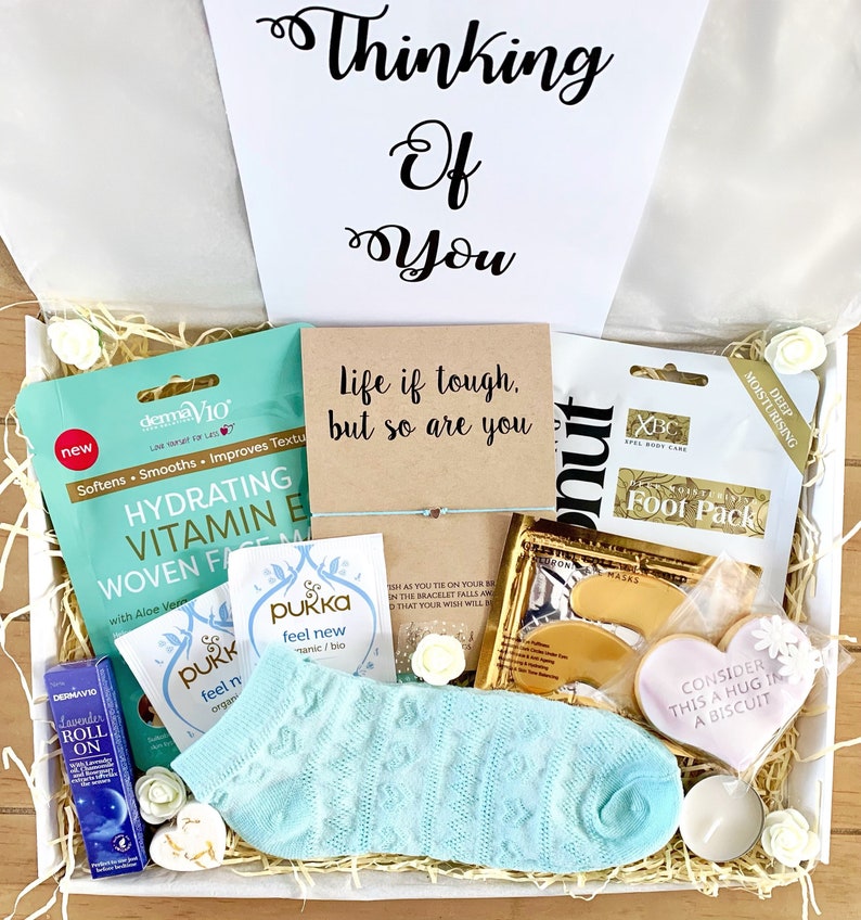 May include: A white gift box filled with self-care items, including a face mask, foot mask, socks, a candle, and a cookie. The box also includes a card with the text "Life if tough, but so are you".